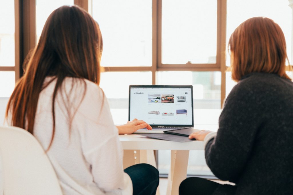 Two people with long hair sit facing away in front of a window, looking at a laptop together.