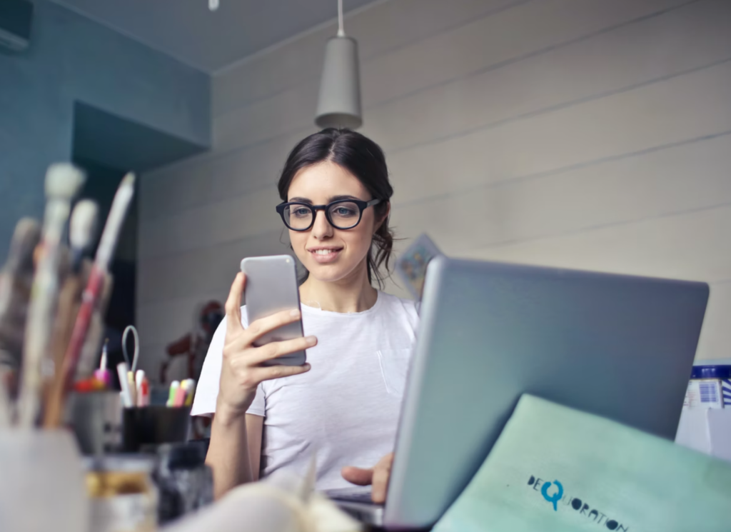 woman wearing glasses is looking at phone, laptop open, money matters at fleming college