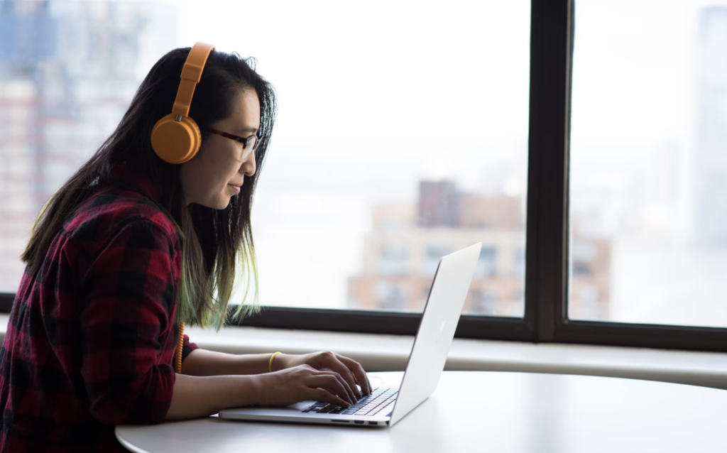 woman leaning over open laptop wearing yellow headphones, money matters at cambrian college