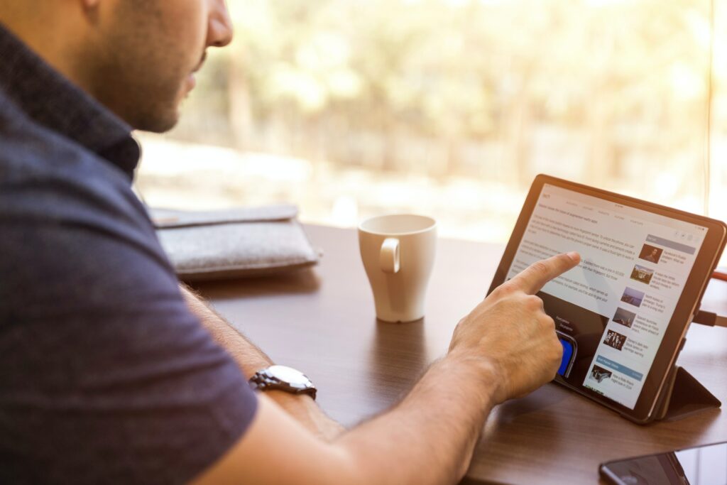 A student sits at a desk in daylight with a cup of coffee, pointing at text on his iPad as he reads.