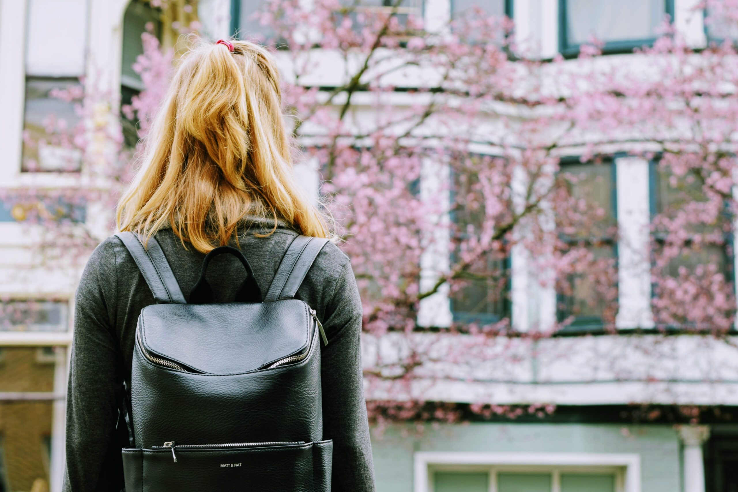 A student with shoulder length blonde hair wearing a backpack looks at a college building.